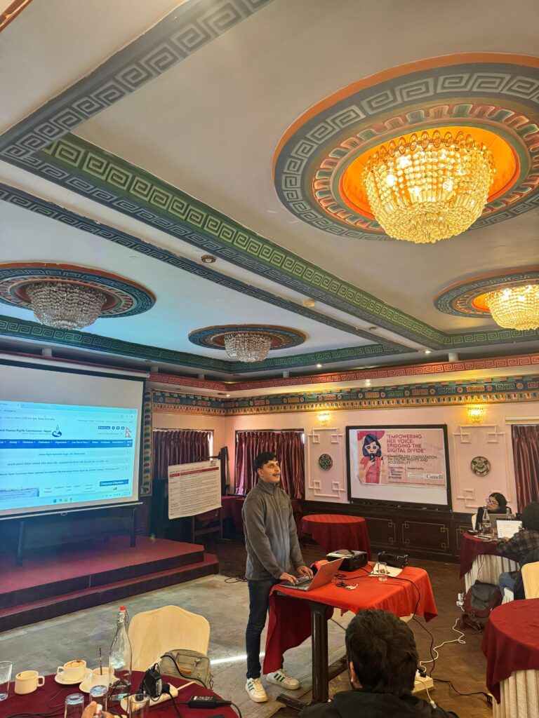 A person stands at a podium in a conference room with ornate ceilings, speaking to an audience. A large screen displays a website, and posters are visible in the background.