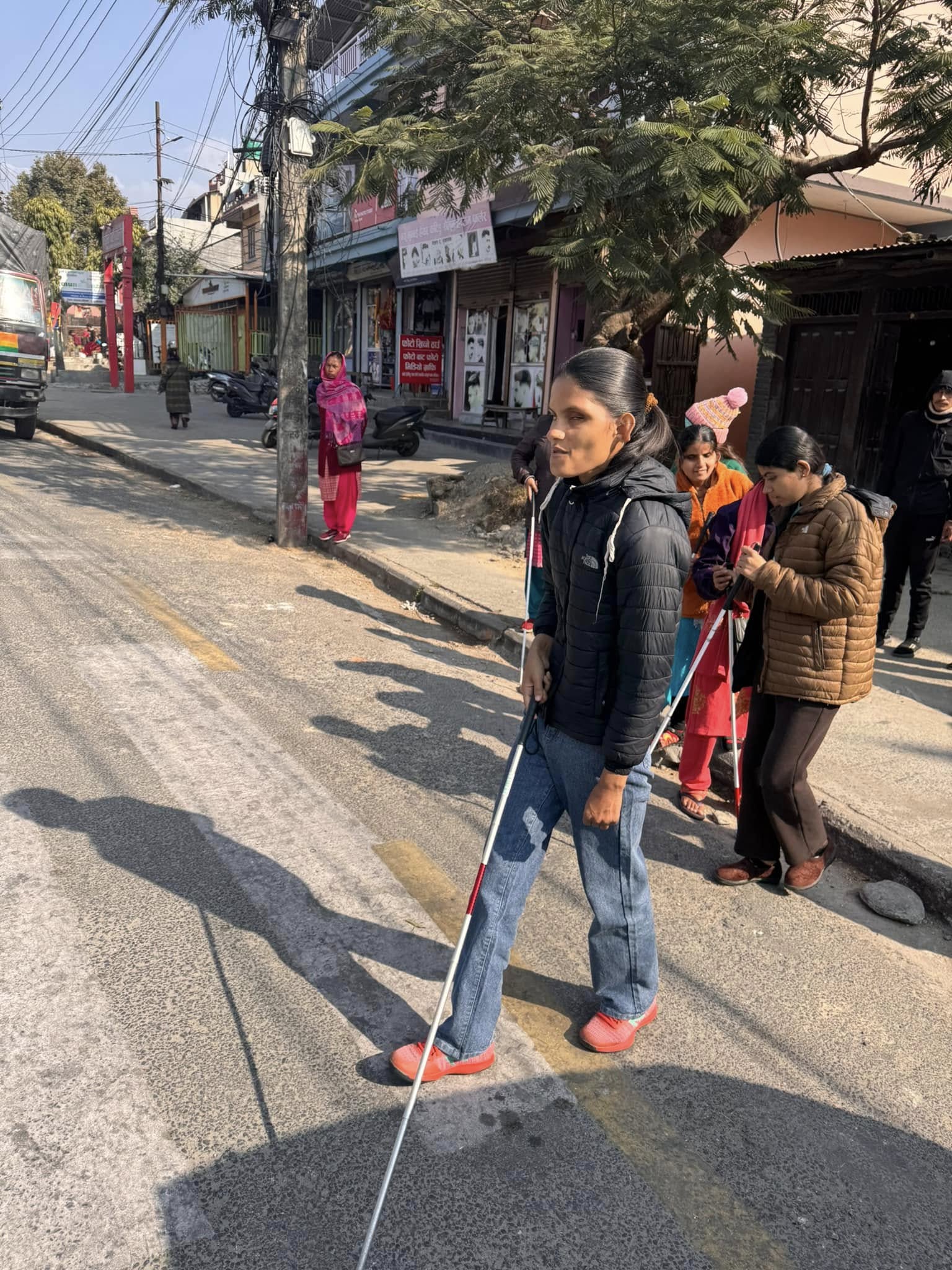 Person with a white cane walking on a sunny street with shops and other pedestrians.