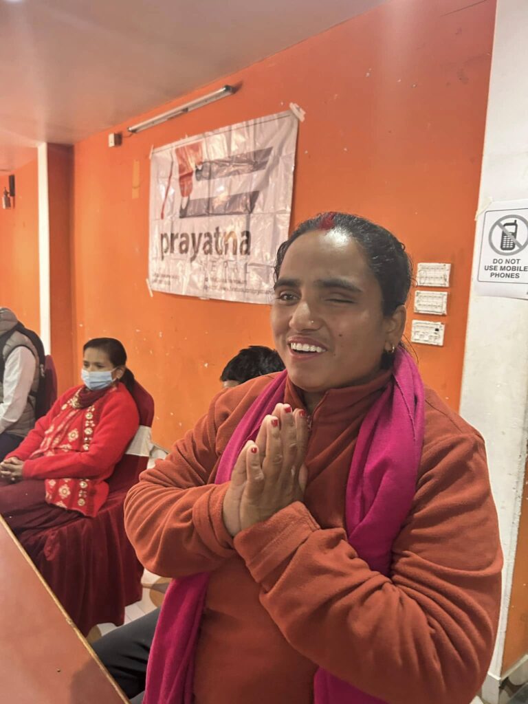 A woman in an orange and pink outfit stands indoors with hands pressed together, smiling. Two people sit in the background near an orange wall and a "Prayatna" banner.